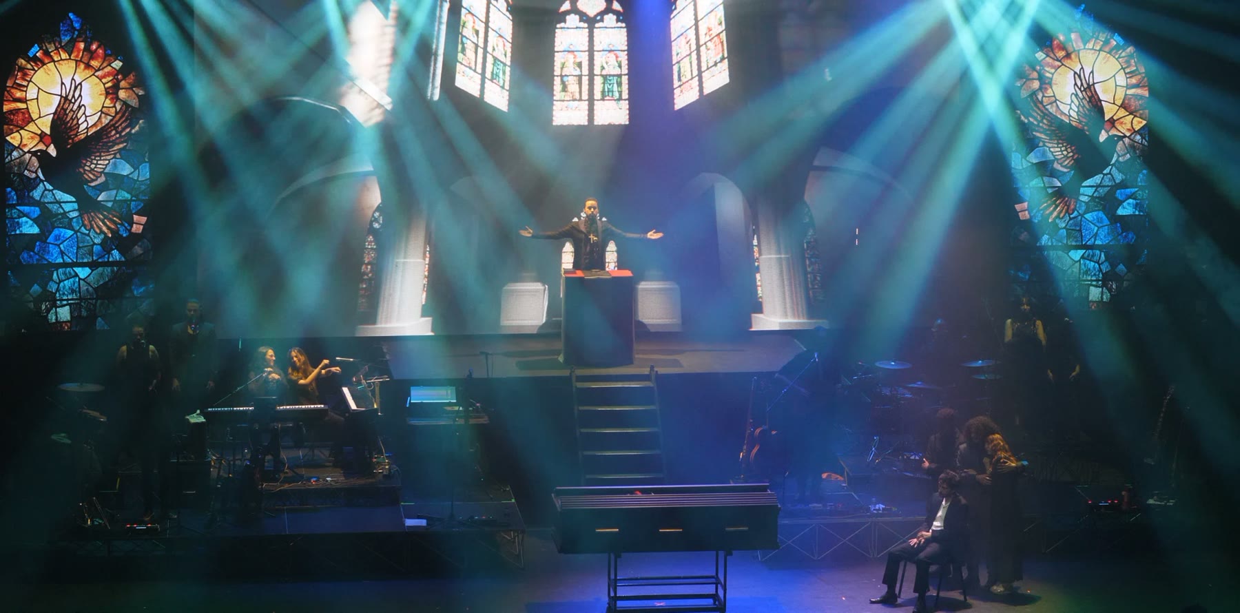 Montaje de gran formato: banda y elenco en escena, con vitrales proyectados y luces cruzadas